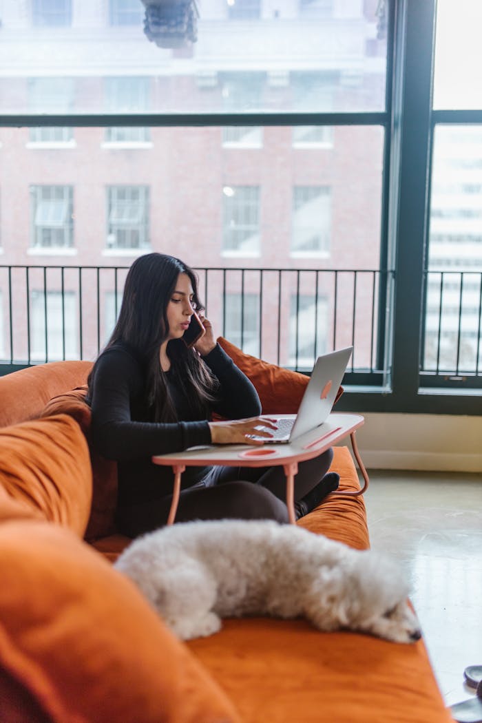 Young woman multitasking with laptop and phone on cozy sofa at home.